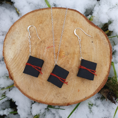 Three miniature books with black book cloth covers, and red ties. Two are on chain drop earrings, one is hung on a necklace. They are artfully displayed on a cut log surrounded by snow and greenery.