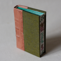 A small book with green fabric cover and tan leather spine, held to show off the bee and flowers hand-painted on the foredge.