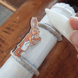 A wooden pin badge in front of a book spine being smoothed with a bonefolder. The badge is a cartoon image of a rabbit in an apron, holding a bonefolder.