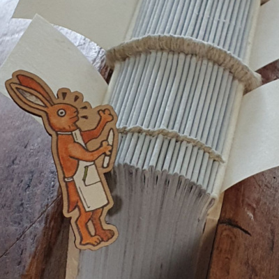 A wooden pin badge in front of a book spine. The badge is a cartoon image of a rabbit in an apron, holding a bonefolder.
