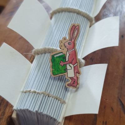 A wooden pin badge in front of a book spine. The badge is a cartoon image of a rabbit in an apron, holding a book.